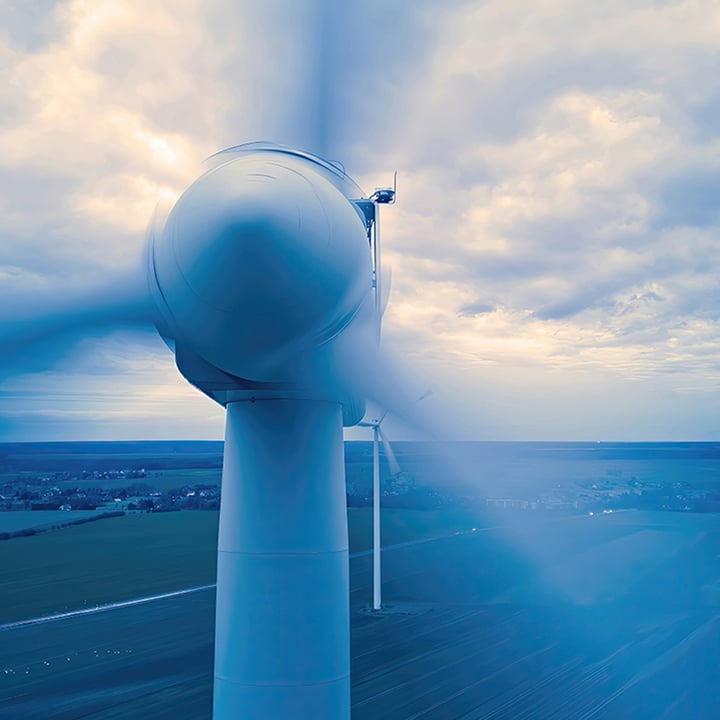 A close-up, low-angle shot of a large white wind turbine with its blades in motion, against a cloudy sky and a faint landscape in the background.