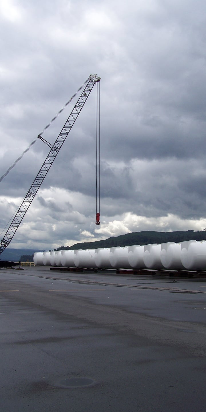 A large crane lifting a component, likely for a wind turbine, at a port with several other components or structures wrapped in white material laid out on the ground, under a cloudy sky